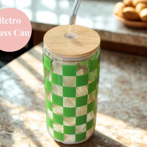 Puede incluir: Una lata de vidrio transparente con un patrón de cuadros verdes y blancos. Tiene una tapa de bambú y una pajita transparente. El texto "Retro Glass Can" está en un círculo rosa. La lata está sobre una superficie clara.