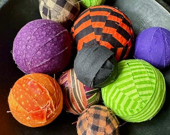 May include: A collection of ten fabric-covered spheres in various sizes and patterns, including black and orange stripes, purple, lime green, orange polka dots, and black and tan buffalo check, all resting together in a dark, round bowl.