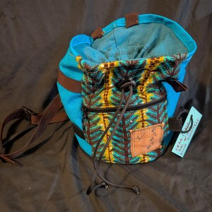 May include: A bright blue chalk bag with a brown and green pine tree pattern on the front pocket. It features a black drawstring closure, a brown adjustable strap, and a small rectangular patch. A light blue tag is attached with the text Crag-Cals.