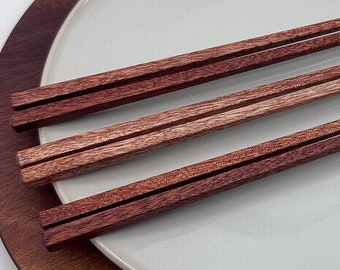 May include: A set of four wooden chopsticks on a brown wooden plate with a white ceramic plate inside. The chopsticks are a reddish brown color.