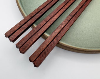 May include: Four pairs of dark red wooden chopsticks on a green ceramic plate. The chopsticks are arranged in a neat stack.