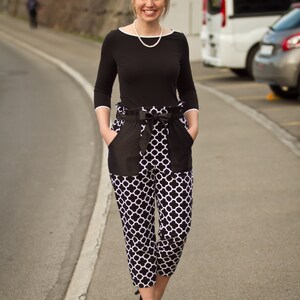May include: A woman wearing a black top with a pearl necklace and black and white patterned pants. She is standing on a paved road with a white line.