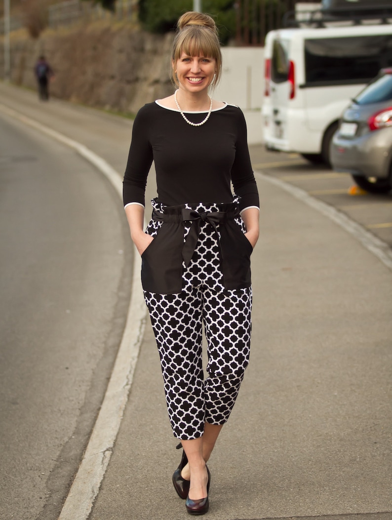 May include: A woman wearing a black top with a pearl necklace and black and white patterned pants. She is standing on a paved road with a white line.