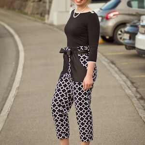 May include: A woman wearing a black top with a white pearl necklace and black and white patterned pants. She is standing on a paved road with cars parked in the background.