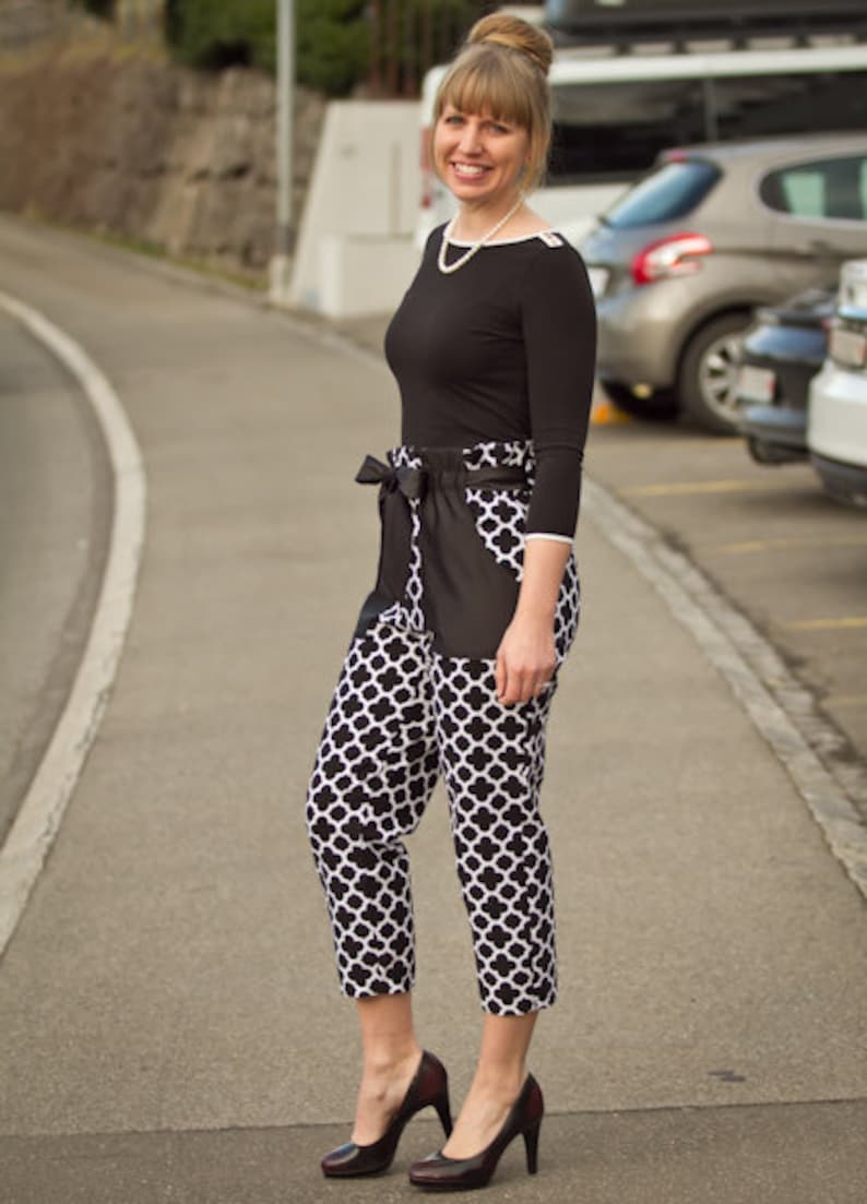 May include: A woman wearing a black top with a white pearl necklace and black and white patterned pants. She is standing on a paved road with cars parked in the background.