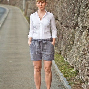 May include: A woman wearing a white button-down shirt and gray shorts with a white belt. She is standing in front of a stone wall.