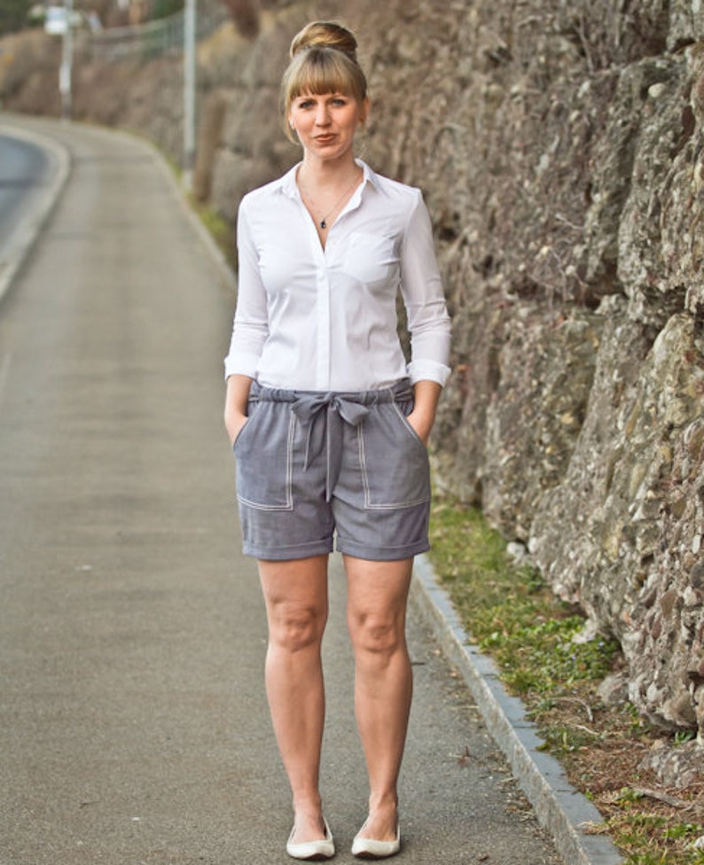 May include: A woman wearing a white button-down shirt and gray shorts with a white belt. She is standing in front of a stone wall.