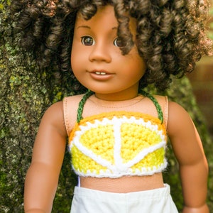 May include: A doll with brown skin and curly brown hair is wearing a yellow and white crocheted top with a green strap. The doll is wearing white shorts and is standing in front of a green mossy tree.