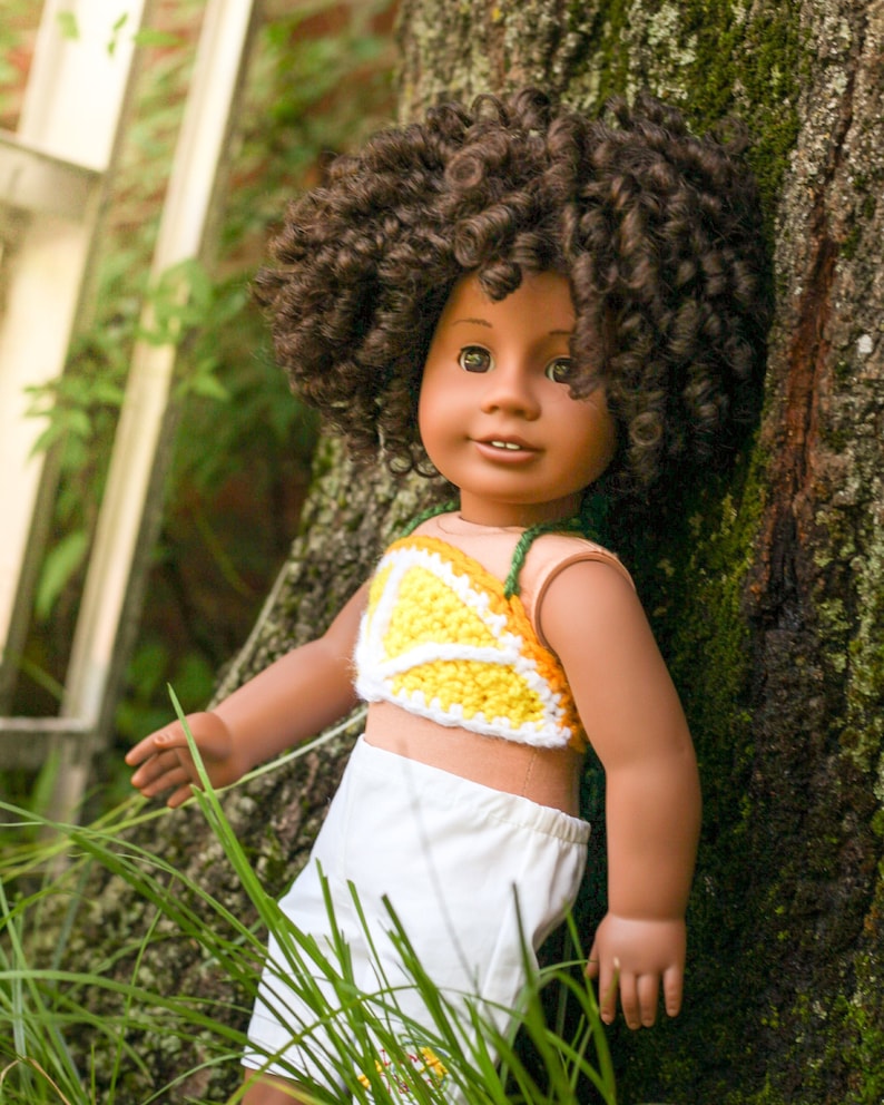 May include: A doll with brown curly hair wearing a yellow and white crocheted top and white shorts. The doll is standing next to a tree.