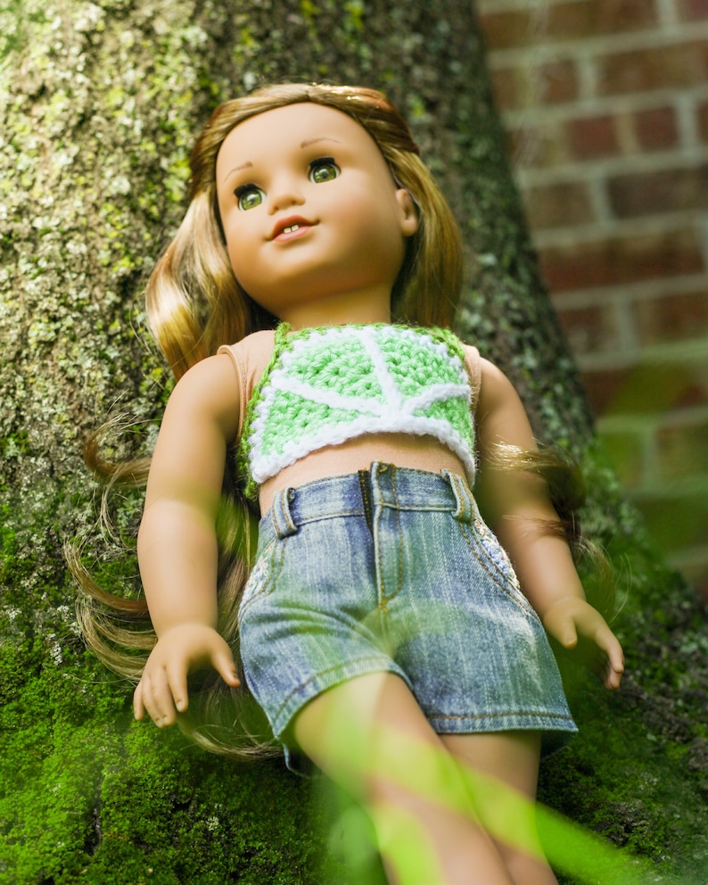 May include: A doll wearing a green and white crocheted halter top and denim shorts. The doll has long brown hair and blue eyes. The doll is leaning against a tree trunk covered in moss.