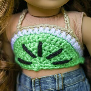 May include: A green crocheted halter top with black stitching, resembling a watermelon. The top is worn by a doll with brown hair and a tan shirt.