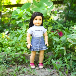 May include: A doll with long brown hair wearing a grey shirt with the text "American Girl" and a denim skirt with a ruffle trim. The doll is standing in a garden setting.