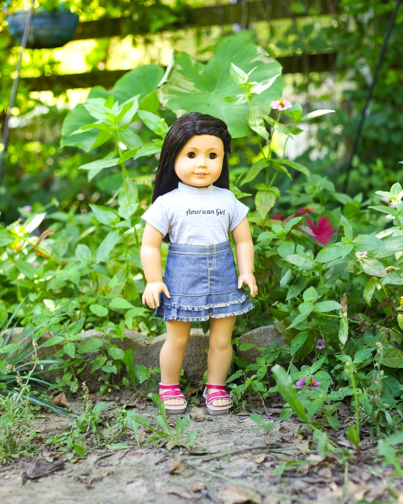 May include: A doll with long brown hair wearing a grey shirt with the text "American Girl" and a denim skirt with a ruffle trim. The doll is standing in a garden setting.