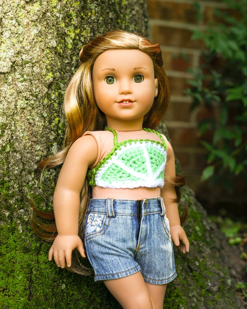 May include: A doll with long brown hair wearing a green and white crocheted lime slice top and blue denim shorts.