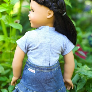 May include: A doll wearing a grey short-sleeved shirt and a blue denim skirt with a frayed hem. The doll has long black hair styled in a braid and a ponytail. The skirt has a red "American Girl" label.