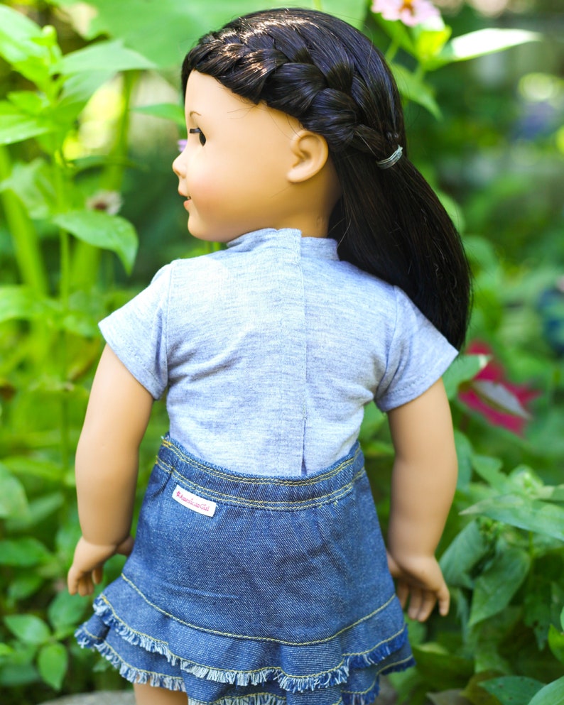 May include: A doll wearing a grey short-sleeved shirt and a blue denim skirt with a frayed hem. The doll has long black hair styled in a braid and a ponytail. The skirt has a red "American Girl" label.
