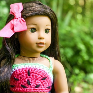 May include: A doll with long brown hair wearing a pink bow, a watermelon-patterned crochet top, and blue jeans. The doll has brown eyes and is looking to the right.