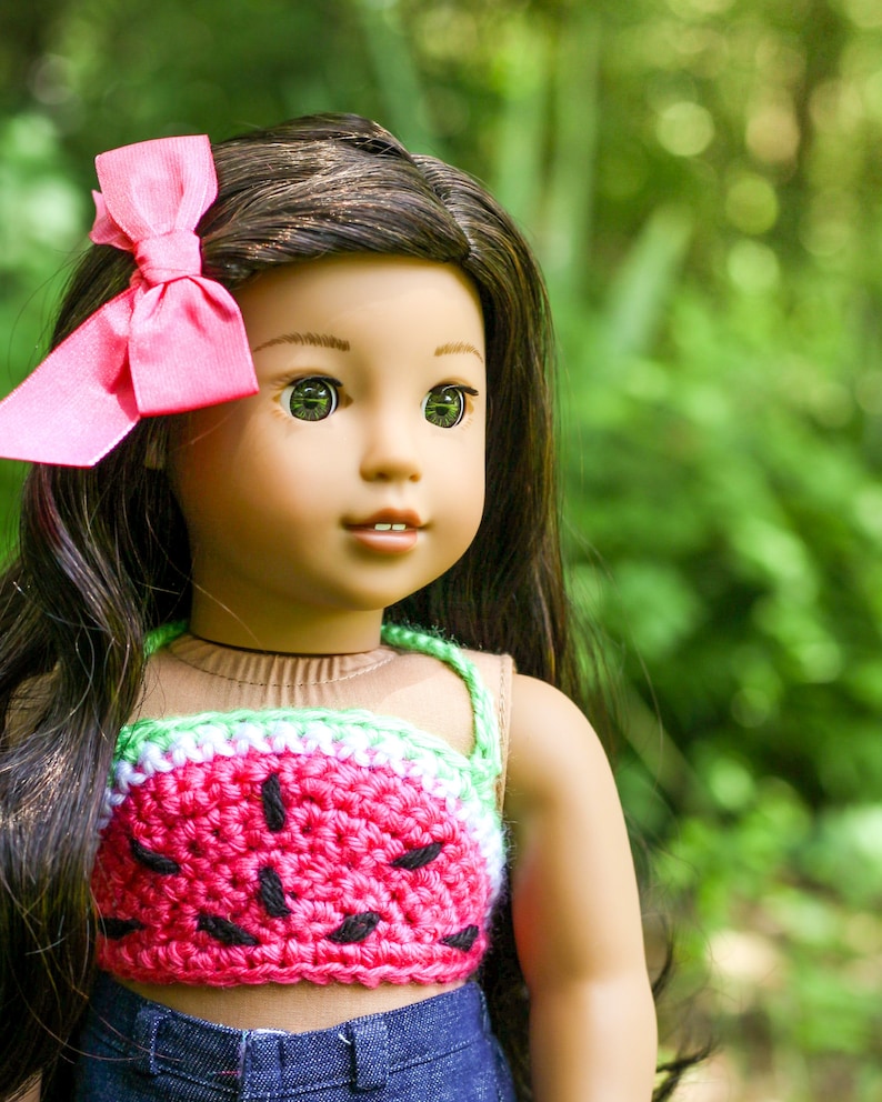 May include: A doll with long brown hair wearing a pink bow, a watermelon-patterned crochet top, and blue jeans. The doll has brown eyes and is looking to the right.