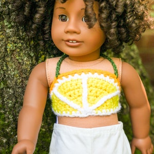 May include: A doll with brown skin and curly brown hair is wearing a yellow and white crocheted crop top with a lemon design. The doll is wearing white pants and is standing next to a tree.