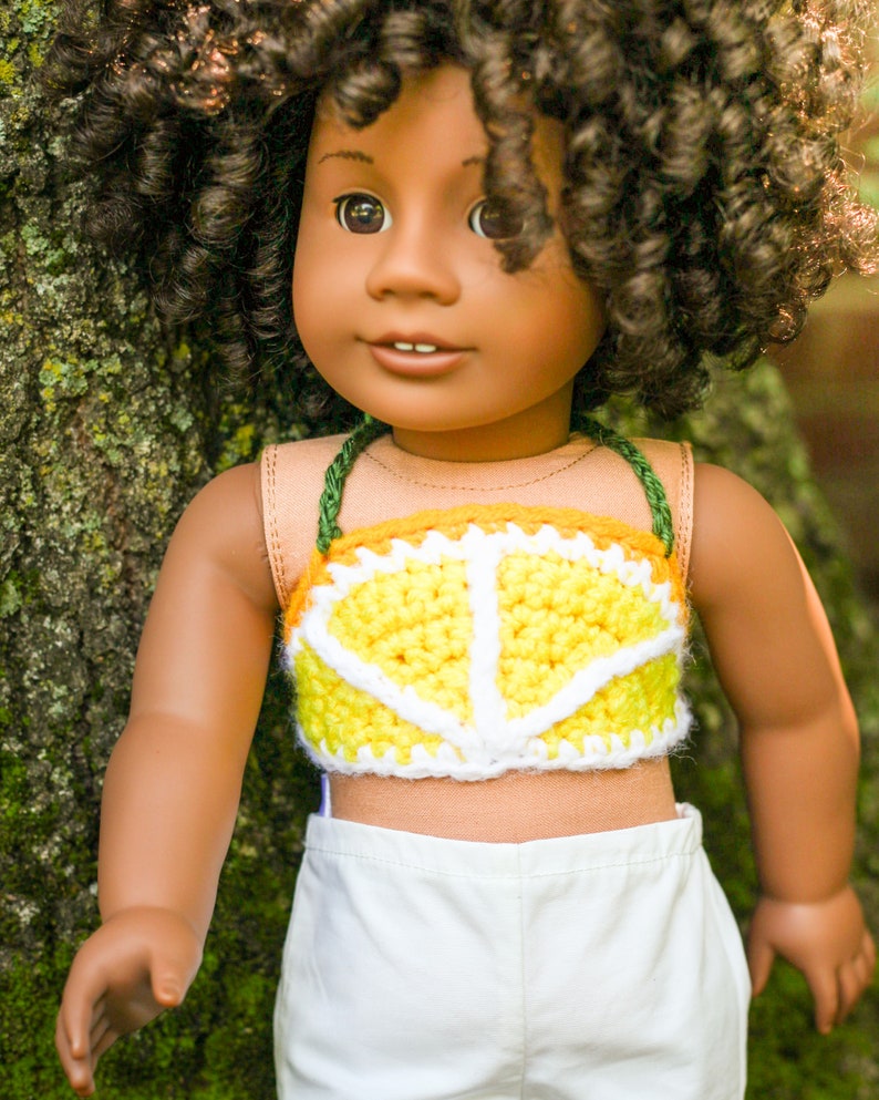 May include: A doll with brown skin and curly brown hair is wearing a yellow and white crocheted crop top with a lemon design. The doll is wearing white pants and is standing next to a tree.