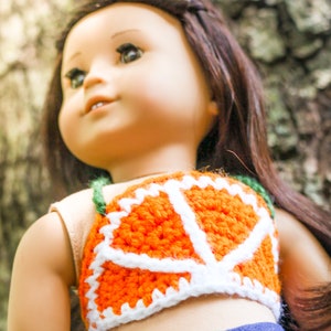 May include: A doll wearing a crocheted orange and white top with a green strap. The top is shaped like a slice of orange. The doll has long brown hair and is wearing blue jeans.