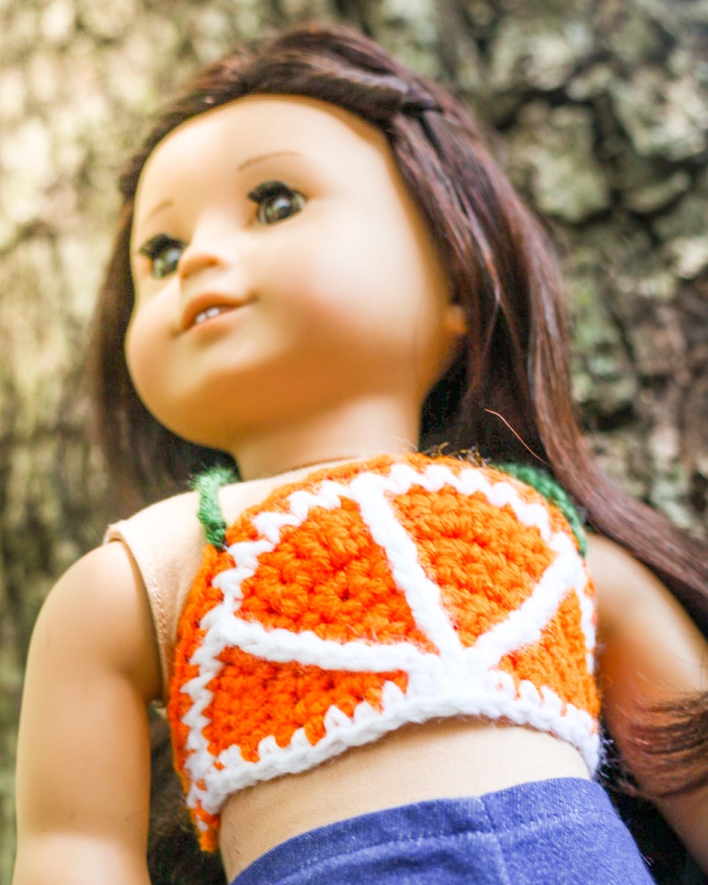 May include: A doll wearing a crocheted orange and white top with a green strap. The top is shaped like a slice of orange. The doll has long brown hair and is wearing blue jeans.