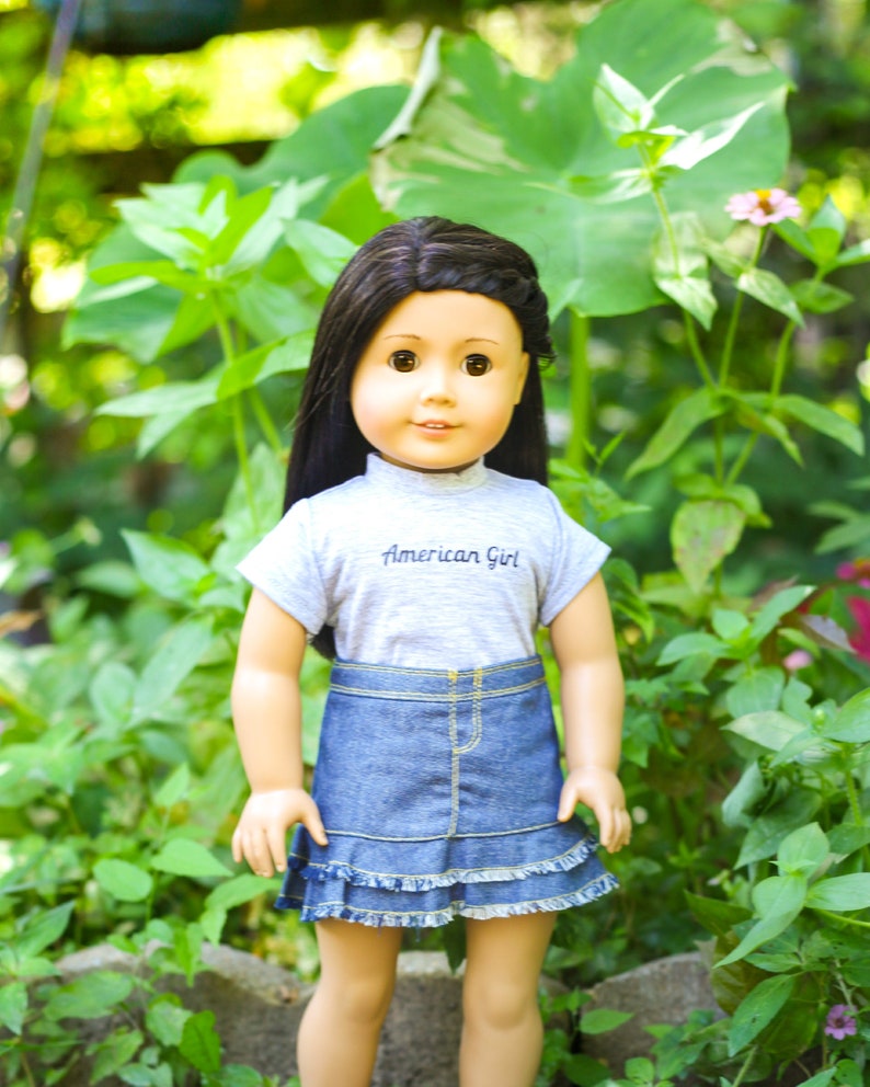 May include: A doll with long dark hair wearing a grey t-shirt with the text "American Girl" and a blue denim skirt with a ruffled hem.