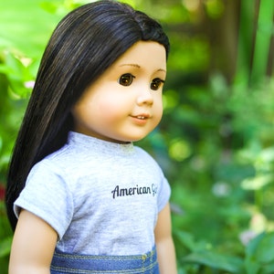 May include: A doll with long black hair wearing a grey t-shirt with the text "American Girl" and blue denim jeans. The doll is looking to the right and has brown eyes.