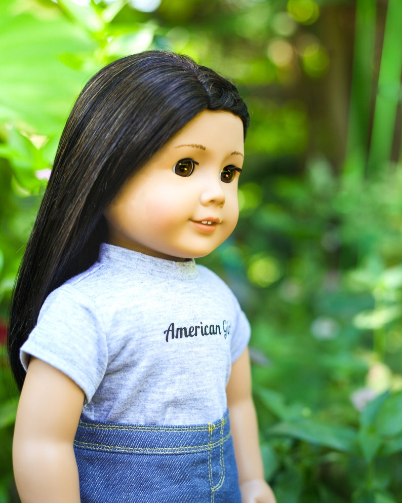 May include: A doll with long black hair wearing a grey t-shirt with the text "American Girl" and blue denim jeans. The doll is looking to the right and has brown eyes.