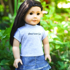 May include: A doll with long black hair wearing a grey t-shirt with the text "American Girl" and a blue denim skirt.