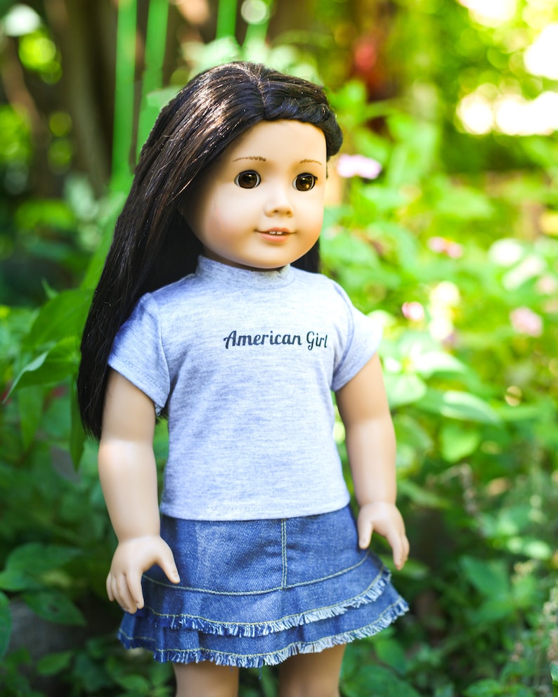 May include: A doll with long black hair wearing a grey t-shirt with the text "American Girl" and a blue denim skirt.