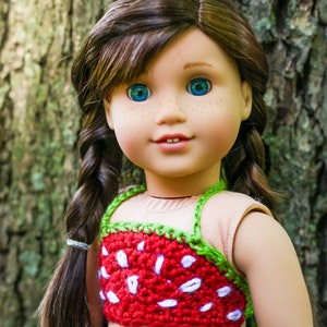 May include: A doll with long brown hair in braids wearing a red and green crocheted strawberry top and blue jeans.