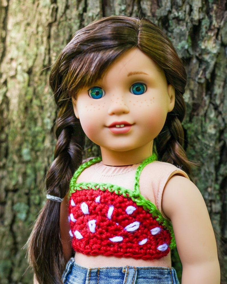 May include: A doll with long brown hair in braids wearing a red and green crocheted strawberry top and blue jeans.
