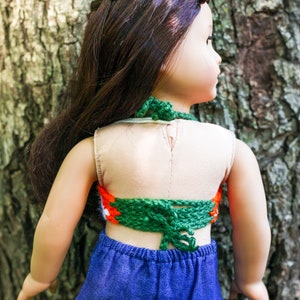 May include: A doll wearing a green, orange, and white crocheted halter top with a blue denim skirt. The doll has long brown hair in a braid.