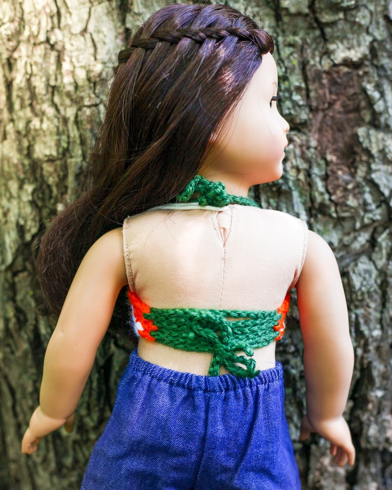 May include: A doll wearing a green, orange, and white crocheted halter top with a blue denim skirt. The doll has long brown hair in a braid.