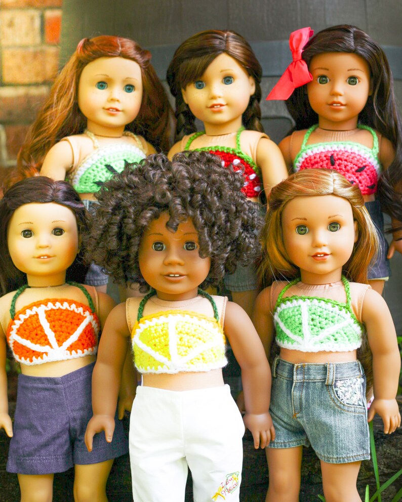 May include: Six dolls wearing crocheted fruit-themed tops. The tops are made with different colors of yarn and feature various fruits, including watermelon, lemons, and oranges. The dolls are wearing different colored shorts and have different hairstyles.