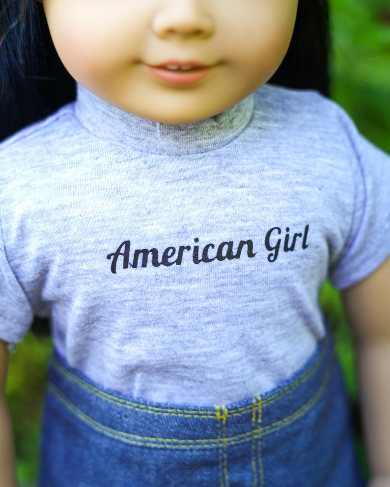 May include: A gray t-shirt with the text "American Girl" printed on the front. The shirt is worn by a doll with long black hair and is paired with blue denim jeans.
