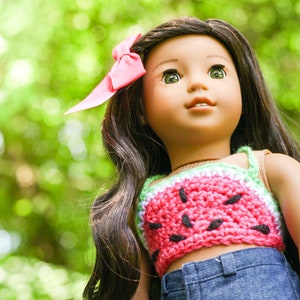 May include: A doll with long dark brown hair wearing a pink and green watermelon crochet top and blue jeans. The doll is looking up and has a pink bow in her hair.