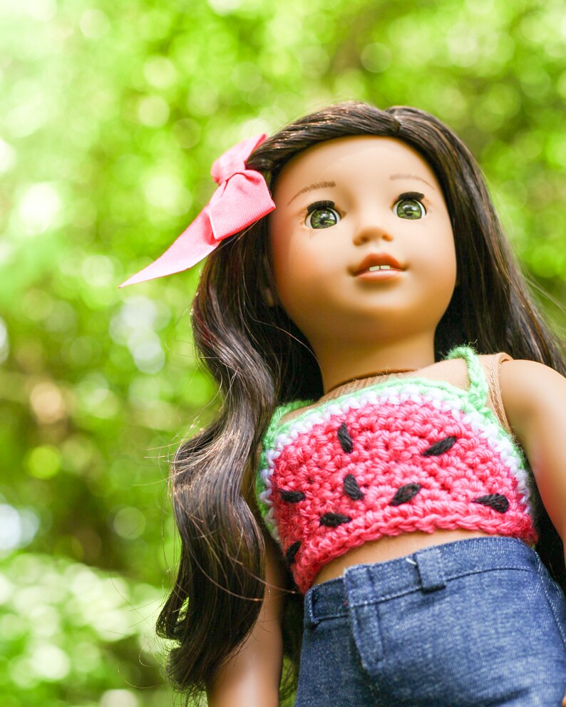 May include: A doll with long dark brown hair wearing a pink and green watermelon crochet top and blue jeans. The doll is looking up and has a pink bow in her hair.