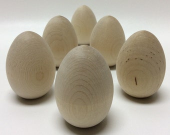 May include: Six unfinished wooden eggs, each with a smooth, rounded surface. The eggs are arranged in a semi-circle on a white background.