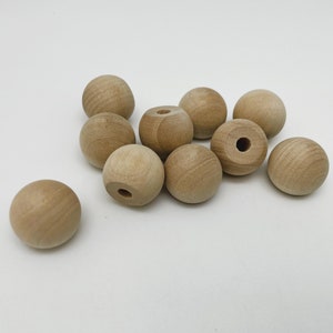 May include: A group of ten natural wood round beads with a hole in the centre. The beads are light brown in colour and are arranged in a cluster on a white surface.