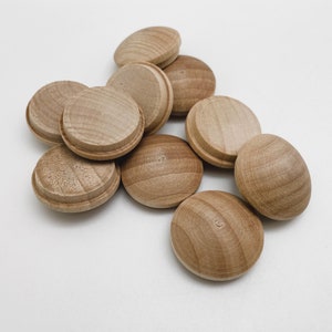 May include: A collection of unfinished wooden plugs, ideal for furniture or craft projects. The plugs are light brown, round, and have a slightly domed top with a grooved edge. They are scattered on a white surface.