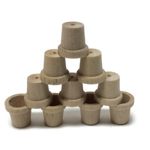 May include: A pyramid of small, unfinished wooden pots. The pots are light brown, with a flared base and a cylindrical top. Each pot has a small hole in the center of the top. The pots are stacked on a white background.