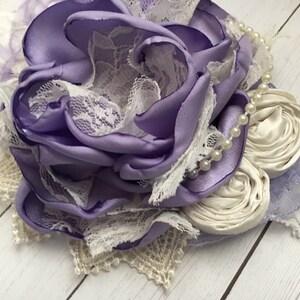 May include: A purple satin flower with white lace and pearl accents. The flower is handmade and would be a beautiful addition to any hair accessory.