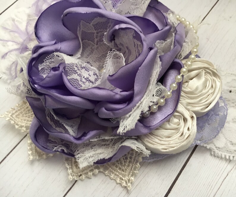 May include: A purple satin flower with white lace and pearl accents. The flower is handmade and would be a beautiful addition to any hair accessory.