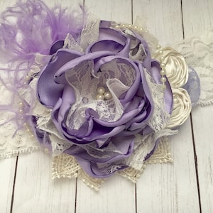 May include: A purple satin flower with white lace and pearl accents. The flower is attached to a white headband with a white feather accent.