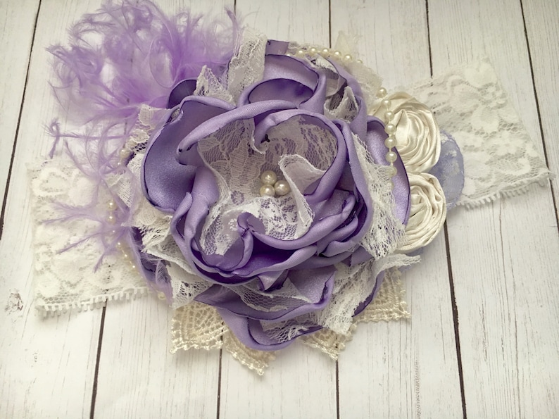 May include: A purple satin flower with white lace and pearl accents. The flower is attached to a white headband with a white feather accent.