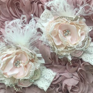 以下が含まれることがあります： 淡いピンク色の布製の花、白いレース、パールアクセントが付いた2つのフローラルヘッドバンド。各ヘッドバンドは、布の花びら、羽、繊細なレースバンドが特徴です。背景は、ダスティローズ色のテクスチャード加工されたフリル生地です。