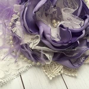 May include: A purple satin flower with white lace and pearl accents. The flower is surrounded by purple feathers.