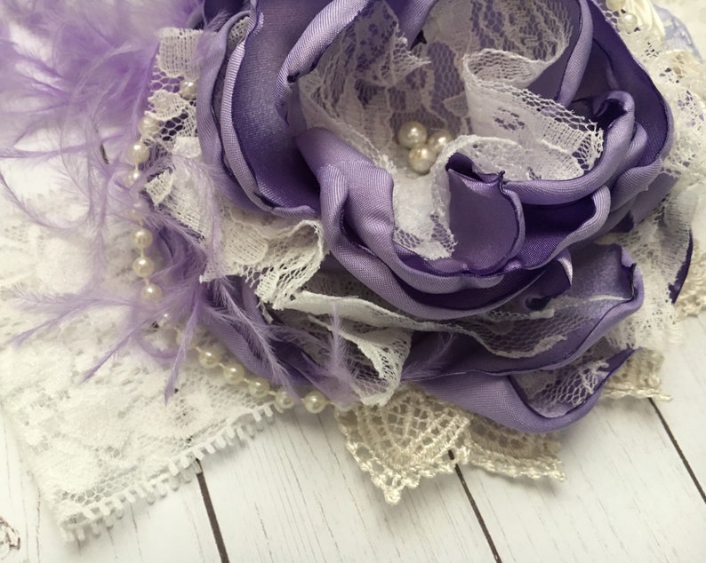 May include: A purple satin flower with white lace and pearl accents. The flower is surrounded by purple feathers.
